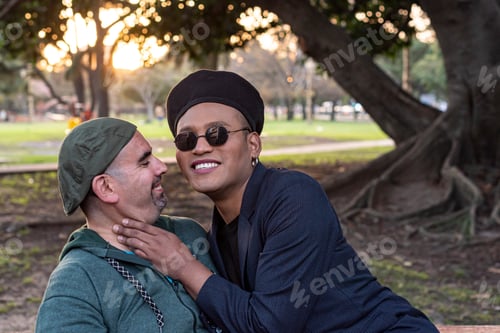Preview: Gay couple hugging each other, talking and laughing while sitting on a beach in a park.