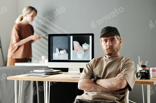 Preview: Young confident photographer in t-shirt and baseball cap sitting by workplace