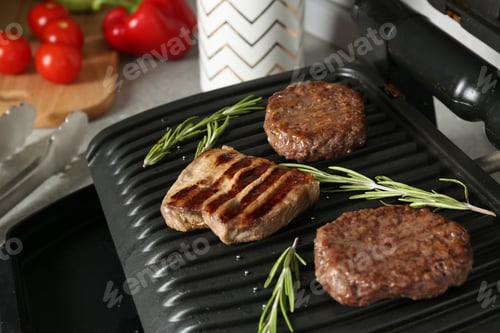 Preview: Open electric grill with beef steak, cutlets and rosemary on table, closeup