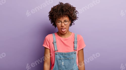 Preview: Photo of young curly woman feels disgust, sticks out tongue, sees something loathsome, has dissatisf