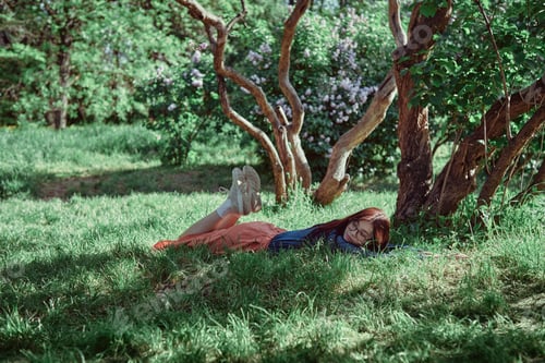 Preview: Teenage girl lying in the grass under a blooming lilac tree, enjoying a serene moment in garden.