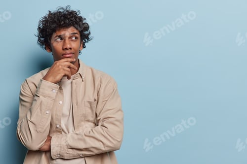 Preview: Horizontal shot of thoughtful curly haired Hindu man keeps hand on chin being deep in thoughts wears