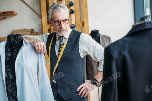 Preview: handsome tailor leaning on mannequin at sewing workshop and looking at jacket