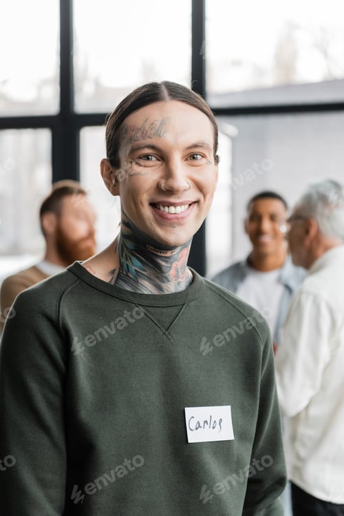 Preview: Portrait of smiling tattooed man with name sticker looking at camera during aa meeting in rehab