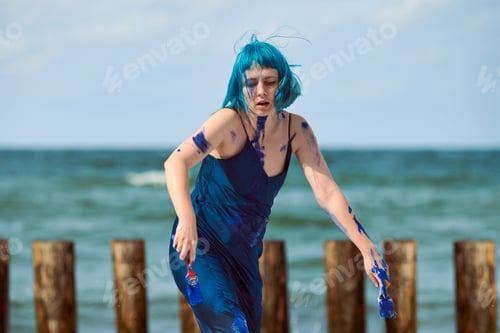 Preview: Artistic blue-haired woman performance artist smeared with blue gouache paints dancing on beach