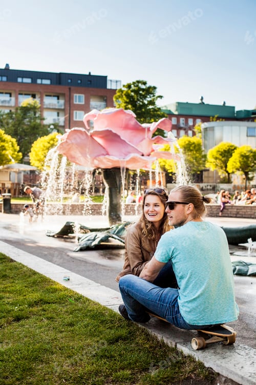 Preview: Happy woman sitting with man on skateboard at park in city