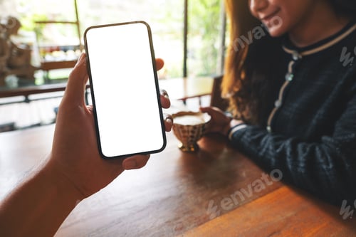 Preview: a man's hand holding black mobile phone with blank white screen with woman drinking coffee in cafe