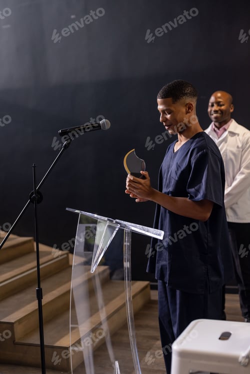 Preview: Young man in scrubs holding award at podium, smiling with colleague nearby