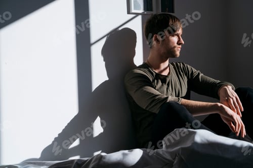 Preview: Serious young man sitting on bed at home