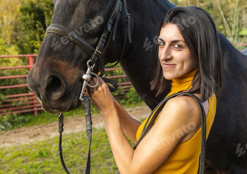 Preview: Woman smiling while adjusting a black horse's bridle. Lifestyle. Horse riding.