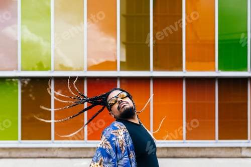 Preview: Latin man tossing his braids back in front of vibrant orange building colorful panels.