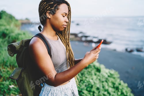 Preview: Dark skinned female tourist with rucksack using internet for networking during trip