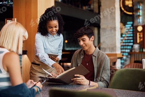 Preview: Happy black waitress recommending order from a menu to her customers in a cafe.