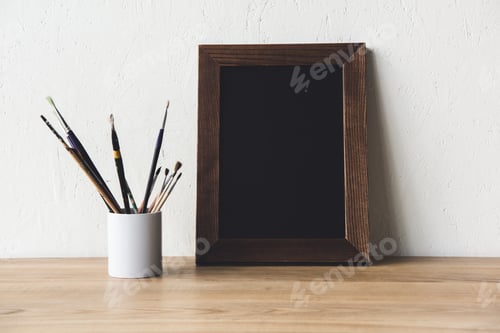 Preview: close up view of empty photo frame and paintbrushes on wooden tabletop
