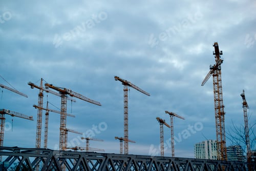 Numerous construction cranes tower against a cloudy sky at a large development site.