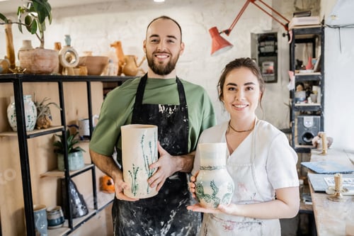 Preview: Smiling couple of artisans in aprons holding clay vases and looking at camera in ceramic workshop
