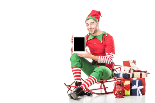 Preview: man in christmas elf costume sitting on sleigh and holding tablet with blank screen isolated on