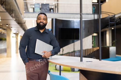 Preview: Business professional holding laptop, standing confidently in modern office lobby, copy space