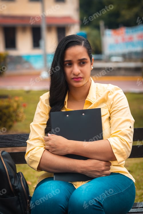Preview: young hispanic college girl holding black folder, sitting in park very worried