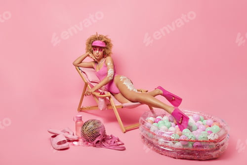 Preview: Dreamy relaxed curly woman keeps lips folded looks aside with thoughtful expression enjoys sunbathin