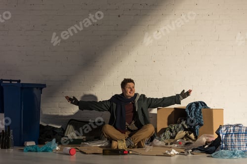 Preview: smiling homeless man sitting on cardboard surrounded by rubbish and spreading arms