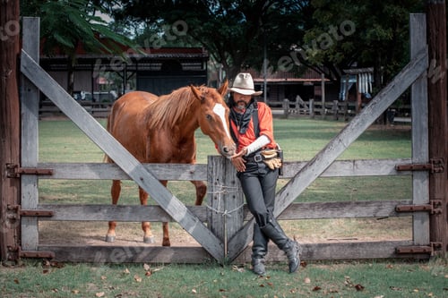 Preview: Portrait cowboy wearing dress vintage style and hat pose taking care and feed his brown horse by lov