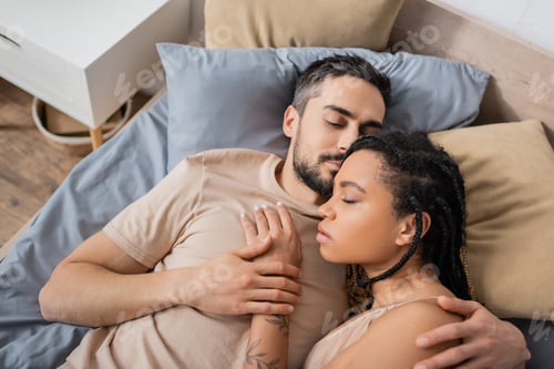 Preview: top view of bearded man with closed eyes hugging sensual african american woman while lying on bed