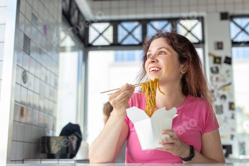 Preview: Portrait of a young cheerful girl eating chinese noodles in a cafe and looking out the window. The