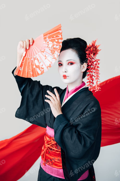 Preview: beautiful geisha in black kimono posing with hand fan and red cloth on background isolated on white