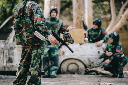 Preview: cropped image of paintball team in camouflage uniform with marker guns resting near broken car