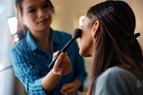 Preview: Close up of Asian makeup artist applying blush on young woman's cheek.
