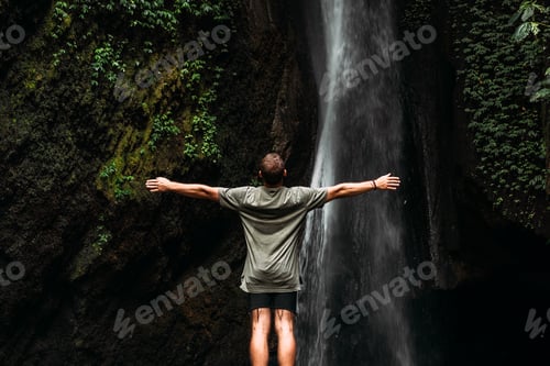 Preview: Back view of a man standing by waterfalls with arms outstretched