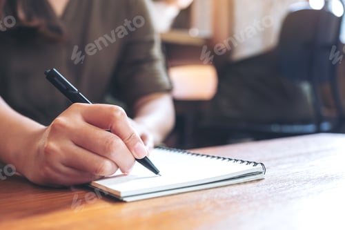 Preview: Closeup image of a woman writing down on a white blank notebook on wooden table