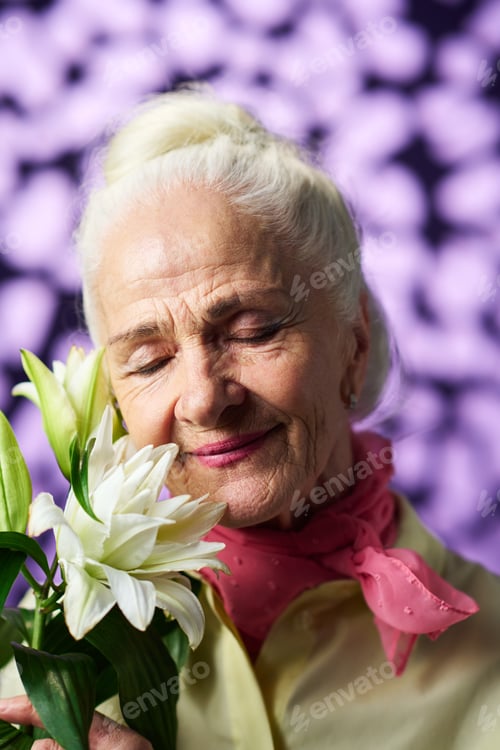 Preview: Happy senior woman enjoying nice smell of gorgeous white lily