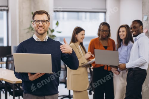 Preview: Professional working on laptop, giving thumbs up with multicultural team collaborating behind