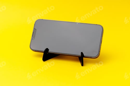 Preview: Smartphone standing on a black smartphone stand isolated on the yellow background