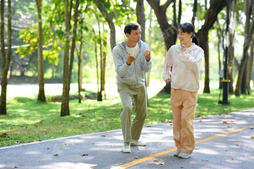 Preview: Cheerful older man and young woman jogging together on a paved path in a beautiful park