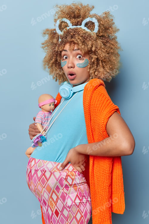 Preview: Shocked pregnant woman stands in profile shows her big belly holds doll anticipates for daughter dre