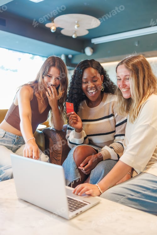 Preview: Diverse young women friends happily buying products online using a laptop and payment card