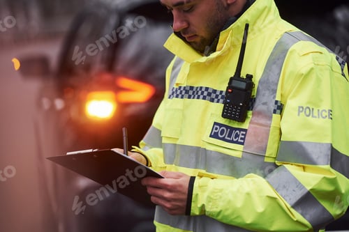 Preview: Male police officer in green uniform standing with notepad near car