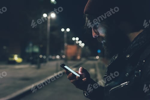 Preview: Young male hipster on city street looking at smartphone at night