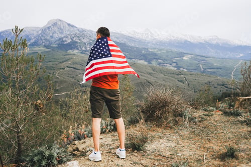 Preview: Young man standing on rock cliff with the US flag on his back while looking at mountains in
