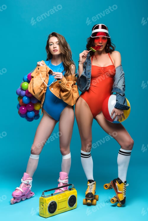 Preview: full length view of two stylish girls in roller skates eating watermelon lollipop on blue