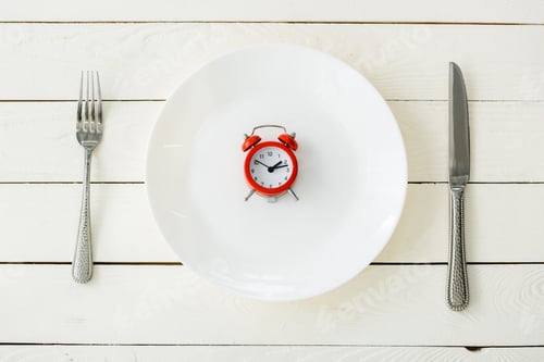 Preview: top view of white plate with red alarm clock near cutlery on wooden surface