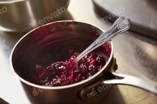 Preview: High angle view of red berry compote in a metal saucepan with a silver spoon.