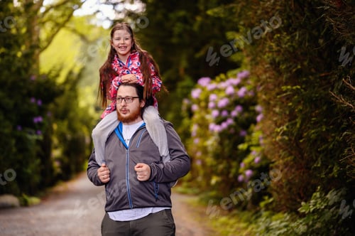 Visualização: Jovem homem barbudo de cabelo comprido em óculos segurando nos ombros menina júnior na estrada no parque