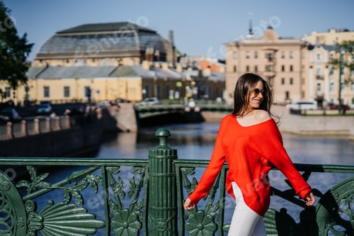 Preview: Cheerful brunette young woman in red sweater and sunglasses walks by bridge against blurry old town