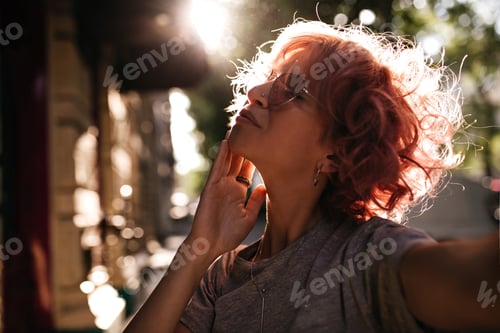 Preview: Thoughtful curly woman in grey tee takes selfie outside. Attractive short-haired girl