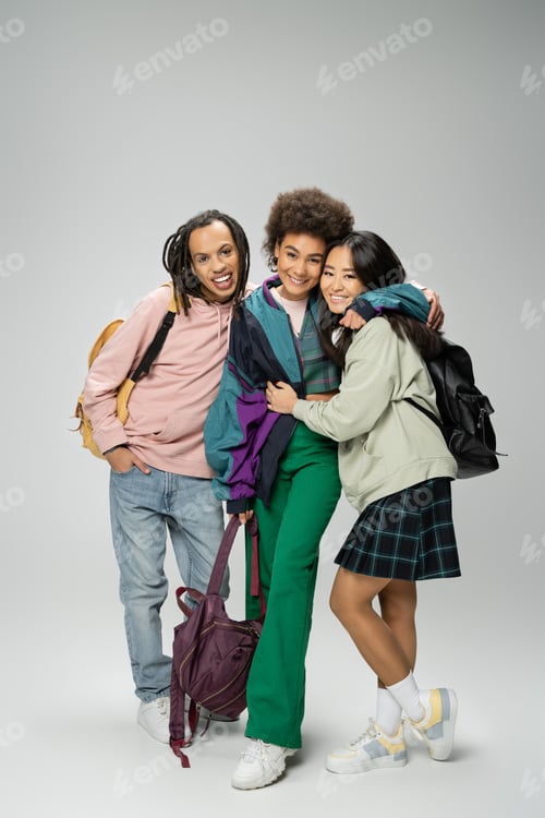 Preview: full length of joyful and stylish multiethnic students with backpacks embracing on grey background