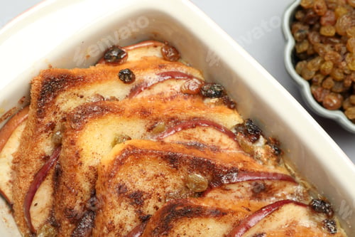 Preview: Delicious bread pudding in baking dish and raisins on light gray table, closeup